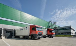 Zwei rote Lkw mit Krone-Fleet-Aufliegern vor einem grün-weißen Logistikgebäude mit Laderampen.
