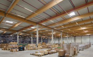 Große Lagerhalle mit Holzbalkendecke, Kartons auf Paletten und Regalen, Betonboden, Tageslichtfenster in der Decke.