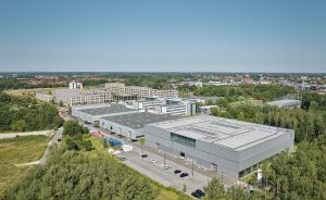 Luftaufnahme eines Industriekomplexes mit grauen Flachdachgebäuden, umgeben von Grünflächen und Bäumen, im Hintergrund eine Stadt.