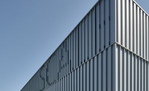 Modernes Industriegebäude mit grauer Metallfassade und vertikalen Lamellen, Glasfront im Erdgeschoss, blauer Himmel.