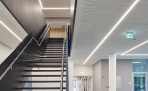 Modernes Gebäudeinneres mit dunkler Stahltreppe, LED-Lichtleisten an Decke, Flur mit Feuerlöscher und Notausgangsschild.