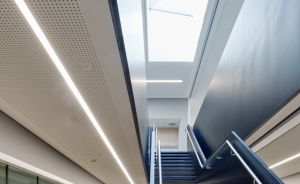 Modernes Gebäudeinneres mit schwarzer Stahltreppe, Oberlichtfenster, linearen LED-Leuchten und Holztheke mit weißen Stühlen.