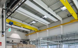 Industriehalle mit gelbem Laufkran an der Decke, Metallregalen und -behältern auf weißem Hallenboden.