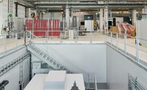 Industriehalle mit Maschinenanlage in einer Grube, Metallgeländer, Treppen, roter Wandverkleidung und Brückenkran an der Decke.