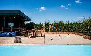 Pool mit blauem Wasser, Holzterrasse, Sitzgelegenheiten, Sandvolleyballfeld mit Netz und Grünhecke im Hintergrund.