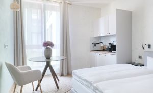 Helles Studio-Apartment mit Doppelbett, kleiner Küche, rundem Glastisch, Stuhl und Balkonzugang.