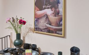 Holzregal mit schwarzem Keramikgeschirr, grüner Vase mit Blumen, dahinter gerahmtes Foto eines Töpfers bei der Arbeit.