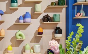 Keramikgeschäft mit bunten Vasen und Schalen auf Holzregalen an rosa Wand; im Vordergrund Teller, Krug und Blumenvase.
