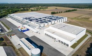 Luftaufnahme eines großen Industriekomplexes mit Solarpanelen auf dem Dach, umgeben von Feldern und Wald.