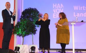 Drei Personen auf einer Bühne, eine hält eine Glasauszeichnung hoch. Im Hintergrund ein "Hannover"-Banner.