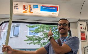 Mann mit Brille und Smartwatch hält sich in einer Straßenbahn in Hannover fest. Im Hintergrund Werbeplakat.