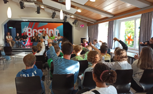 Kinder sitzen in einem Saal und strecken die Hand auf. Auf einer Bühne mit "#Bestjob"-Banner sitzen mehrere Erwachsene.