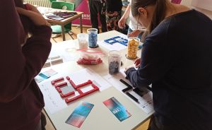 Frau beugt sich über Tisch mit bunten Buchstabenformen aus Legosteinen, Behältern und Zetteln. Weitere Personen stehen dabei.