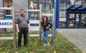 Zwei Personen stehen vor einem Gebäude mit "COMNET IHR SYSTEMHAUS"-Schild auf einer Rasenfläche mit Herbstlaub.