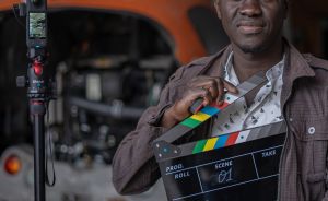 Mann hält Filmklappe (Scene 01) vor Kamera auf Stativ, im Hintergrund ein orangefarbenes Fahrzeug.