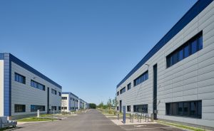 Zwei moderne Industriegebäude aus grauem Metall mit blauen Akzenten, dazwischen ein leerer Asphaltweg, blauer Himmel.