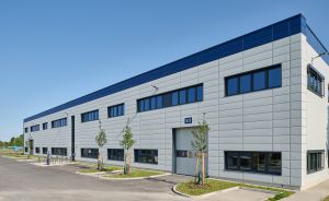 Modernes zweistöckiges Industriegebäude mit grauer Fassade, blauen Akzenten, Nummer 53, Parkplatz und jungen Bäumen.