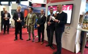 Mehrere Männer in Anzügen stehen vor einem Messestand mit der Aufschrift „German Cities & Regions". Ein Mann spricht ins Mikrofon.