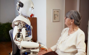 Ältere Frau mit Brille sitzt einem humanoiden Roboter gegenüber und berührt dessen Hand an einem Tisch.