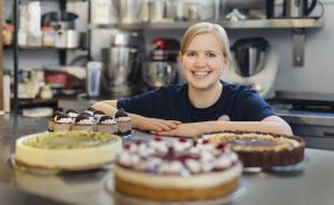 Blonde Frau lächelt hinter mehreren Torten in einer professionellen Bäckereiküche mit Küchengeräten im Hintergrund.