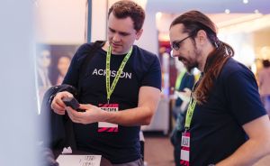 Zwei Männer mit Lanyards schauen gemeinsam auf ein Gerät in einer Hand, im Hintergrund eine Veranstaltung.