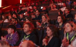 Publikum sitzt in einem abgedunkelten Theater mit roten Sitzen und klatscht; einige tragen grüne Lanyards.