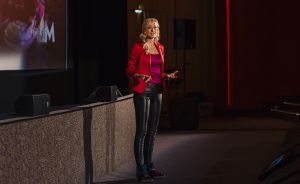 Blonde Frau in rotem Blazer und schwarzer Lederhose spricht auf einer Bühne vor einer Leinwand.