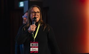 Frau mit Brille und schwarzem Oberteil spricht in Mikrofon. Grünes Lanyard mit rotem „UNLEASH"-Badge sichtbar.