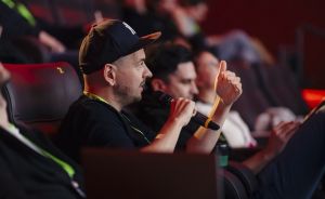Mann mit schwarzer Kappe und Mikrofon sitzt in einem Auditorium, hebt einen Finger und spricht.