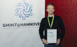 Mann hält Urkunde „1. Platz Publikumsvoting, Nova AI GmbH" vor SMINT@Hannover-Banner mit grünem Lanyard.