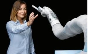 Frau in blauem Hemd interagiert mit einem weißen Roboterarm, der einen Stift hält. Schwarzer Hintergrund.