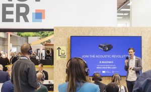 Messestand in Hannover: Presenter vor blauem Bildschirm mit „Join the Acoustic Revolution – Resoniks"-Schriftzug, Publikum im Vordergrund.