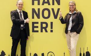 Zwei Personen stehen vor gelbem "We Are Hannover!"-Schild mit Stadtsilhouette und zeigen Daumen hoch.
