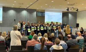 Gruppe von Personen auf einer Bühne hält grüne Zertifikate, davor sitzendes Publikum in einem Konferenzsaal.