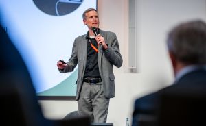 Mann im grauen Anzug mit Mikrofon hält Vortrag vor Publikum, im Hintergrund eine Präsentationsfolie.