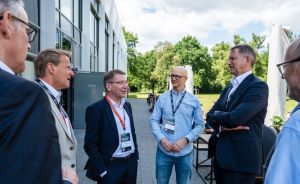 Fünf Männer in Businesskleidung mit Konferenz-Badges stehen im Gespräch vor einem modernen Gebäude im Freien.
