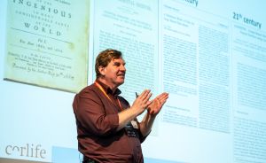 Mann in rotem Hemd spricht vor Leinwand mit historischen Texten und "21st century"-Überschrift; Corlife-Logo sichtbar.