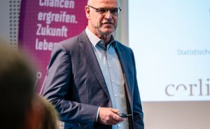 Mann mit Brille und Blazer präsentiert vor Publikum, im Hintergrund Banner mit Aufschrift „DHCH" und Projektionsfläche.