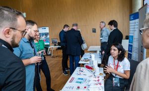 Messestand mit Ausstellerin im Gespräch mit Besuchern; Infomaterialien auf Tisch, Rollups im Hintergrund.