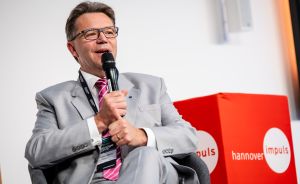 Mann im grauen Anzug spricht in Mikrofon bei Hannover Impuls Veranstaltung, trägt Namensschild und Brille.