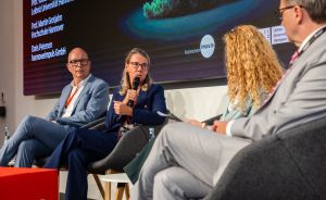 Podiumsdiskussion mit vier Personen; eine Frau spricht ins Mikrofon. Im Hintergrund Namensschilder auf einer Leinwand.