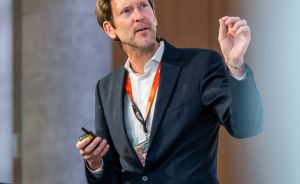 Mann im dunklen Anzug mit orangem Lanyard hält Presenter und gestikuliert an einem Stehpult.