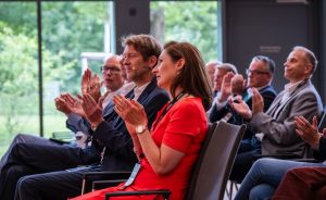 Mehrere Personen in einem Konferenzraum applaudieren sitzend. Eine Frau in Rot trägt ein Namensschild.