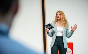 Frau mit blonden Locken hält Vortrag, trägt hellblaues Sakko, hält Heft in der Hand, roter Würfel im Hintergrund.