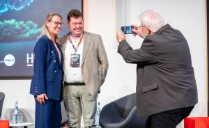 Drei Personen auf einer Bühne: zwei posieren lächelnd für ein Foto, eine dritte fotografiert sie mit einem Smartphone.