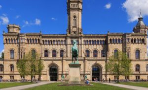 Historisches Gebäude mit Turm und Bogenfenstern, davor grüne Rasenfläche mit bronzenem Reiterstandbild, blauer Himmel.