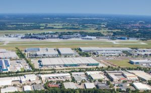 Luftaufnahme eines Industriegebiets mit Lagerhallen neben einem Flughafen, umgeben von Grünflächen und Stadtrand.