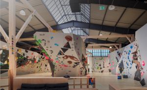 Indoor-Kletterhalle mit bunten Griffen an weißen Boulderwänden, Holzbänken, Skylights und einer kletternden Person rechts.