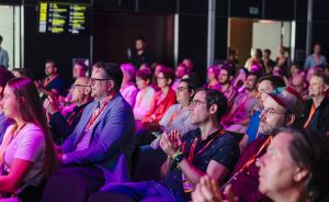 Publikum sitzt in Reihen bei einer Veranstaltung, beleuchtet in Pink/Lila. Mehrere Personen tragen orangefarbene Lanyards.