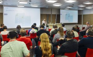 Vortrag in modernem Konferenzraum: Publikum mit Kopfhörern sitzt vor zwei Leinwänden, Redner steht vorne mit Mikrofon.